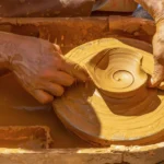 Les mains d'un potier expérimenté accompagnent celles d'un apprenti dans le façonnage de l'argile, illustrant la transmission d'un savoir-faire.